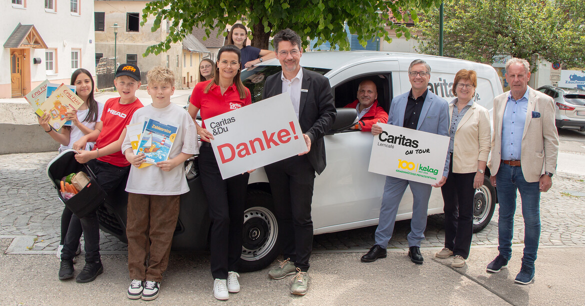 Eine Gruppe von Erwachsenen und Kindern steht im Freien vor einem weißen Fahrzeug. Mehrere Personen halten Tafeln mit der Aufschrift „Danke“ und „Caritas & Du“. 