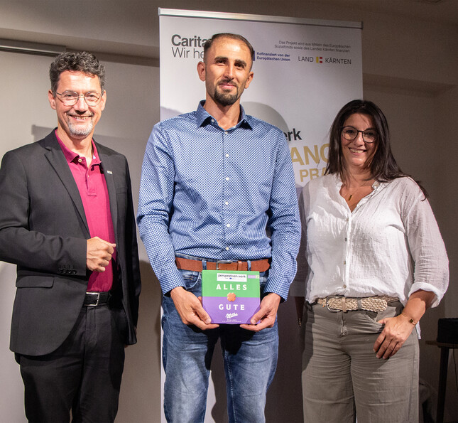 Drei Personen stehen auf einer Bühne, eine Person hält eine Auszeichnung aus dem Arbeitsprojekt