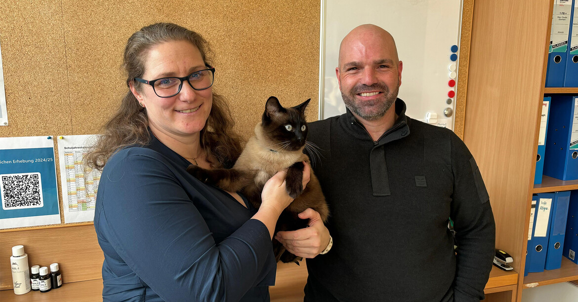 Therapiekater wird von zwei Personen in einem Klassenraum gehalten und unterstützt den pädagogischen Alltag.