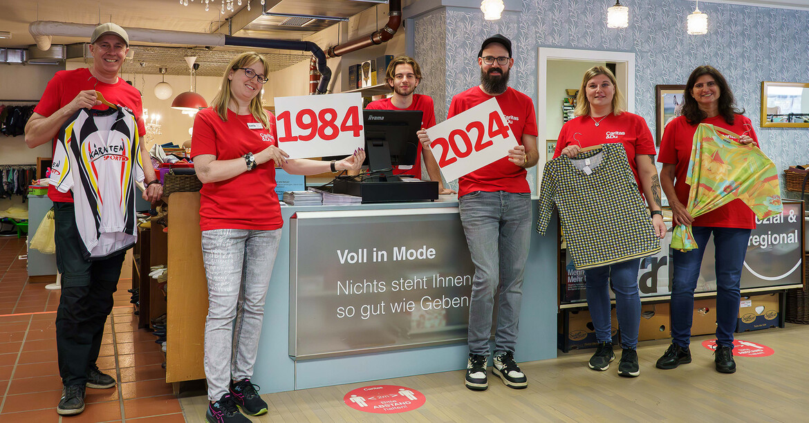 Die Mitarbeiter*innen des carla halten Kleidungsstücke und ein Bild mit der Jahreszahl 1984 und 2024 in den Händen, vor der Kassa im carla Klagenfurt.