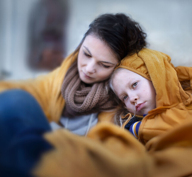 Eine Mutter hat den Kopf auf ihrem Kind abgelegt. Beide sind in einer Decke eingehüllt.