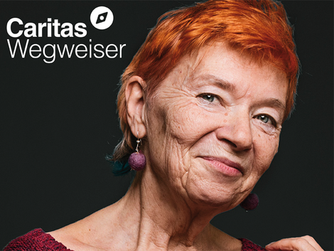 Das Porträt einer Frau ist zu sehen auf schwarzem Hintergrund. Es ist eine ältere Dame die lächelt, sie hat einen rothaarigen Kurzhaarschnitt. Links oben das Logo vom Caritas Wegweiser.