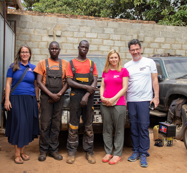 Caritasdirektor Ernst Sandriesser und Projektreferentin Bettina Zangl-Jagiello stehen mit zwei afrikanischen Mechanikern vor ihrer Werkstatt.