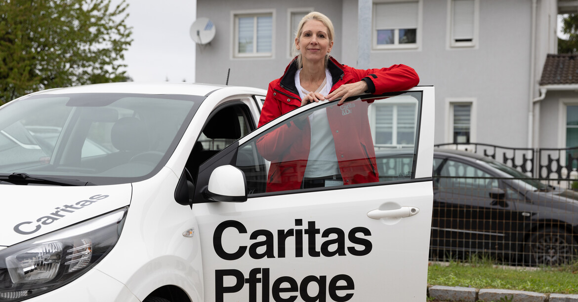 Eine mobile Pflegekraft steht vor der geöffneten Autotür des Dienstautos auf dem Caritas Pflege zu lesen ist.