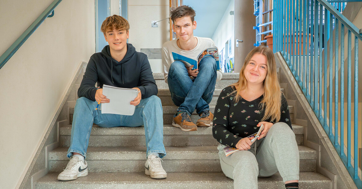 Zwei Schüler und eine Schülerin sitzen auf eine Stiege im Schulgebäude und lächeln.