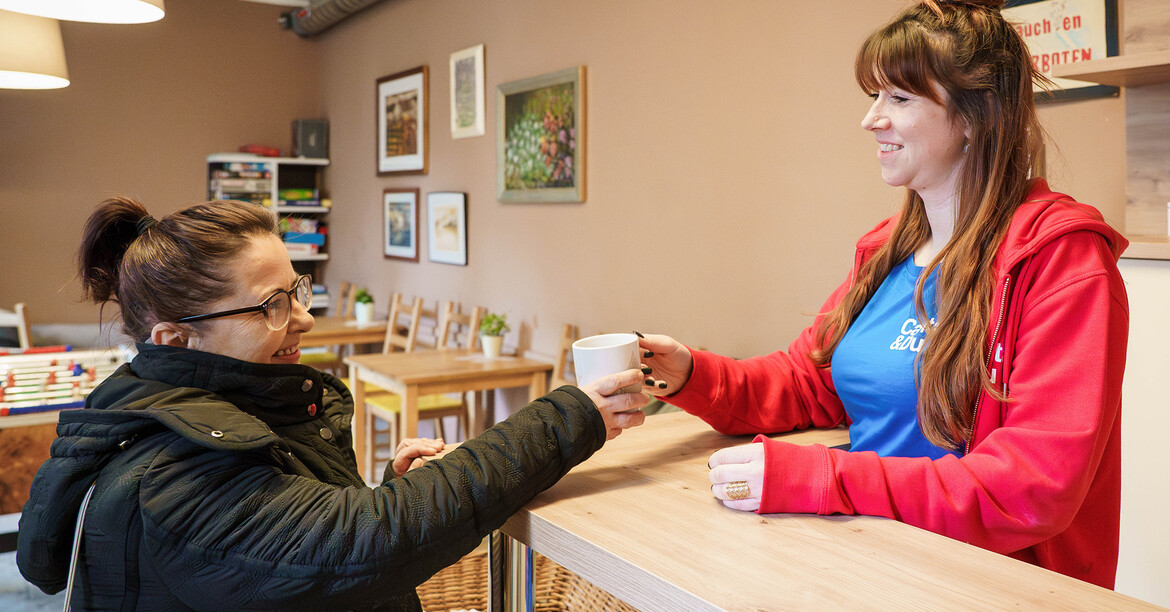 Zwei erwachsene Personen stehen an einer Theke in einem Gemeinschaftsraum. Eine Person hinter der Theke reicht der anderen eine Tasse.