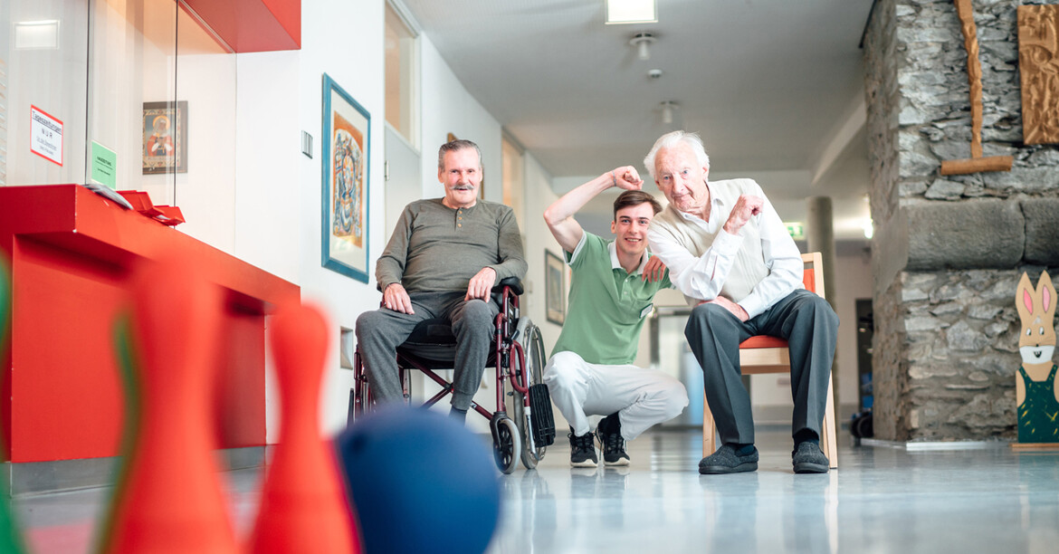 Zivildiener unterstützt zwei ältere Personen bei einer Freizeitaktivität mit Kegeln in einer Caritas-Einrichtung.