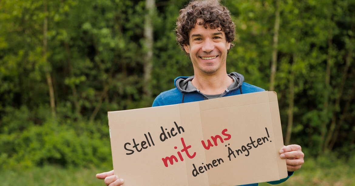 Ein Berater der Caritas-Lebensberatung steht vor grünen Sträuchern und hält ein Schild in der Hand "Stell dich mit uns deinen Ängsten".