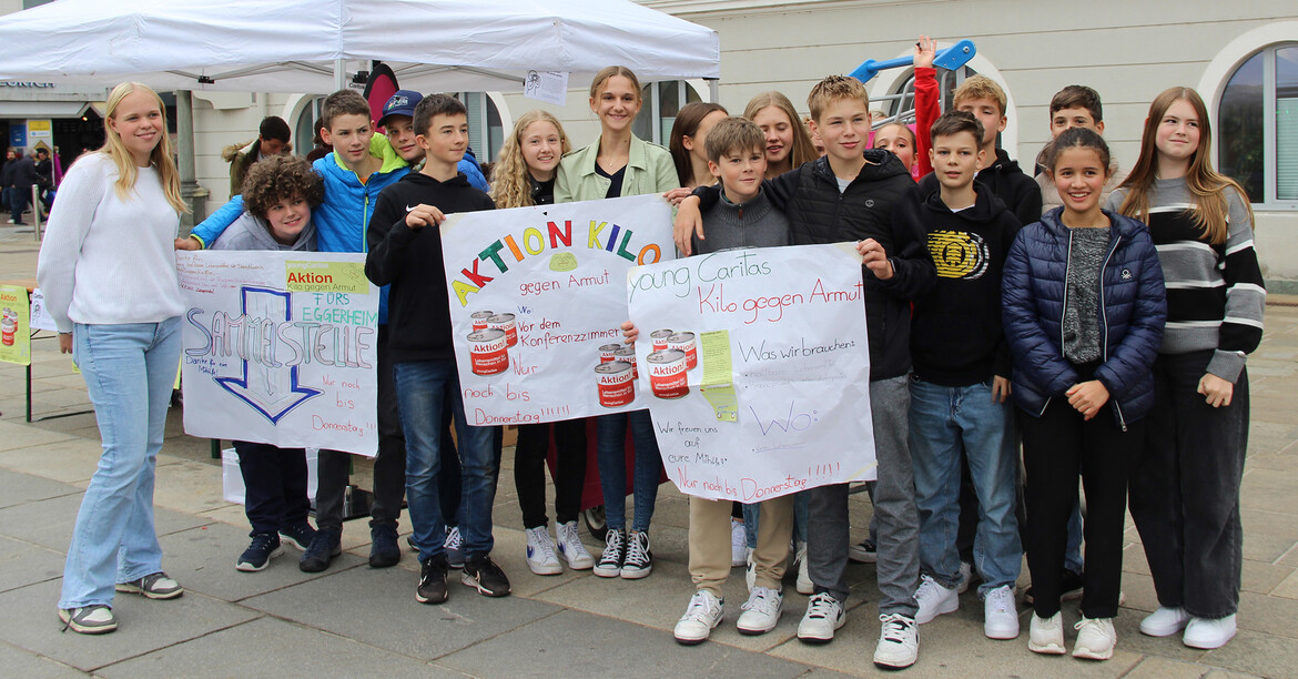 Eine Schülergruppe steht vor der Sammelstation der Aktion Kilo vor dem Rathaus in Klagenfurt. Die Schüler*innen halten Plakate mit Aufschriften zur Aktion in den Händen.