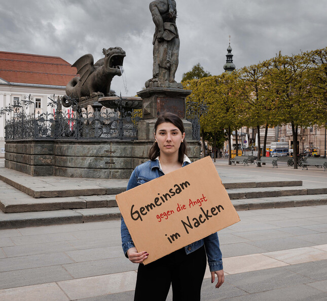 Eine junge Frau steht vor dem Klagenfurter Lindwurm und hält ein Schild in der Hand "Gemeinsam gegen die Angst im Nacken".