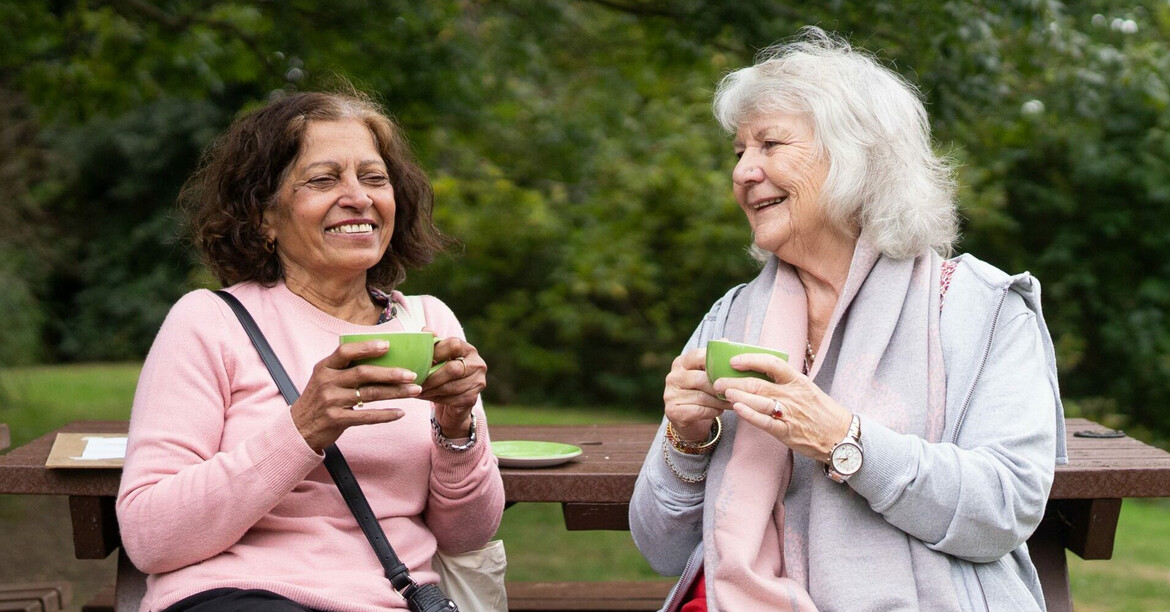 Zwei Frauen sitzen auf einer Picknick-Bank, trinken aus grünen Tassen und lachen miteinander