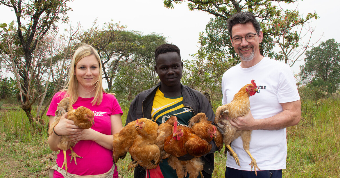 Projektreferentin Bettina Zangl-Jagiello, Caritas-Direktor Ernst Sandriesser und ein Afrikaner halten Hühner in den Händen, hinter ihnen grüne Vegetation.