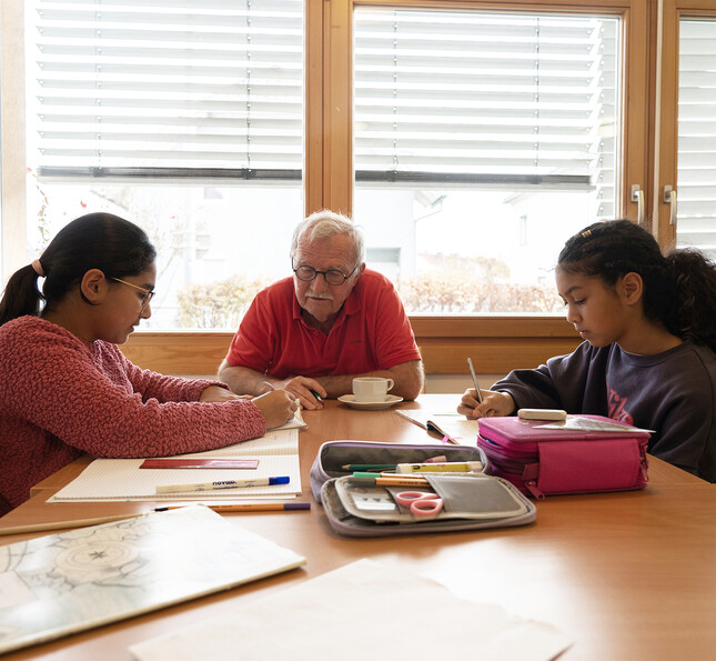 Eine erwachsene Person sitzt gemeinsam mit zwei Kindern an einem Holztisch in einem hellen Raum. 