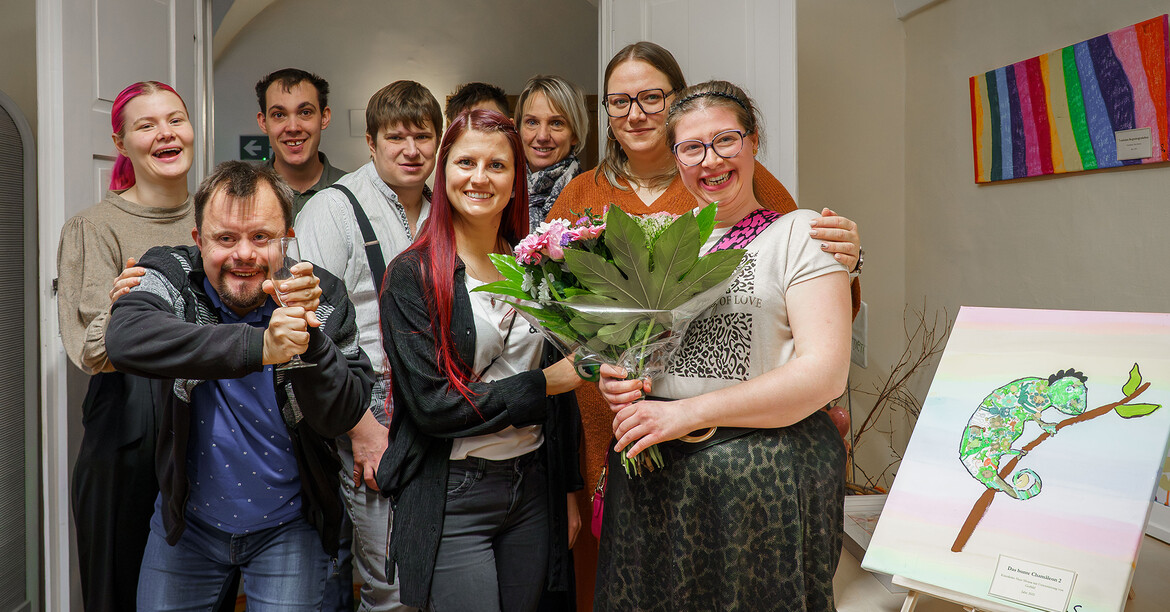 Eine Künstlerin hält einen Blumenstrauß, umgeben von Gästen bei einer Vernissage.