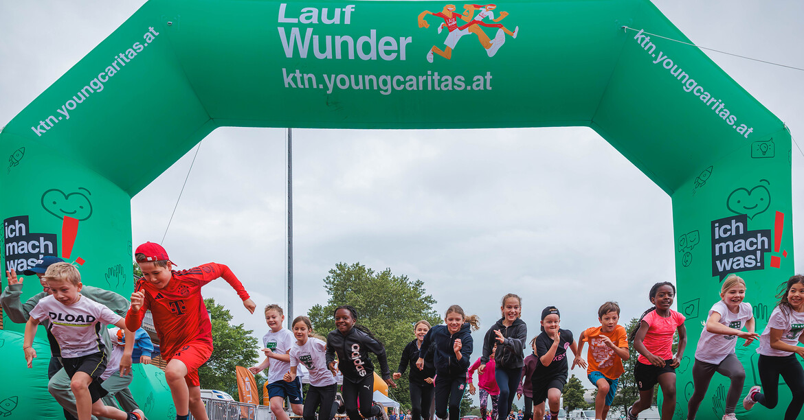 Viele Kinder laufen gemeinsam beim youngCaritas LaufWunder unter einem grünen Startbogen im Freien.