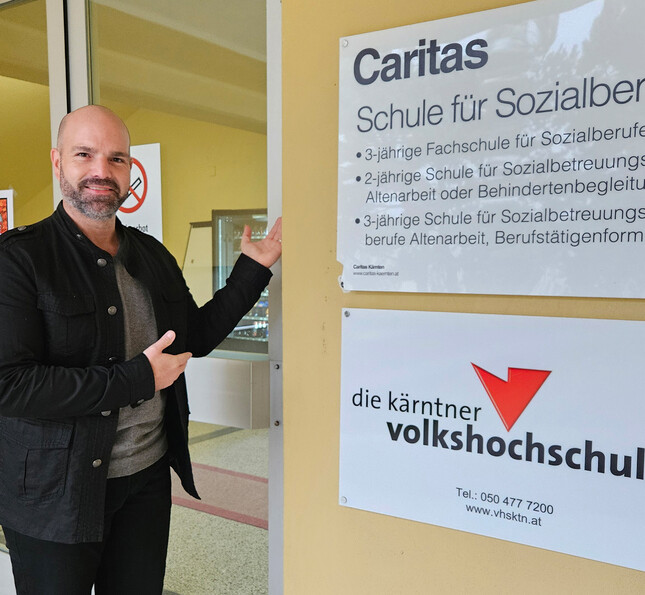 Wolfgang Kogler, Direktor der Schulen für Sozialberufe in Wolfsberg, steht vor der Schule und zeigt auf das Schild mit unseren Ausbildungsmöglichkeiten.