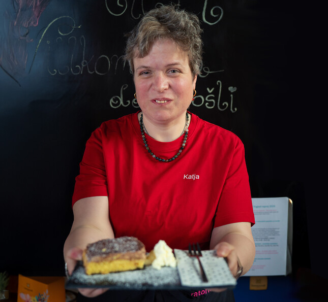 Eine Klientin des Caritas.Team Lebensgestaltung im roten T-Shirt hält einen Teller mit einem Kuchen in der Hand und lächelt.