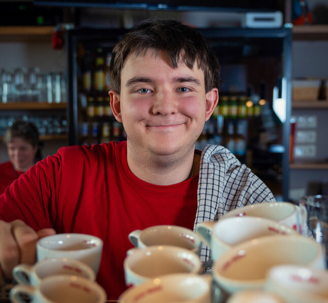 Ein Klient des Caritas.Team Lebengestaltung lächelt in die Kamera, über die Schulter hängt ein Tuch und vor ihm stehen viele Kaffeetassen.