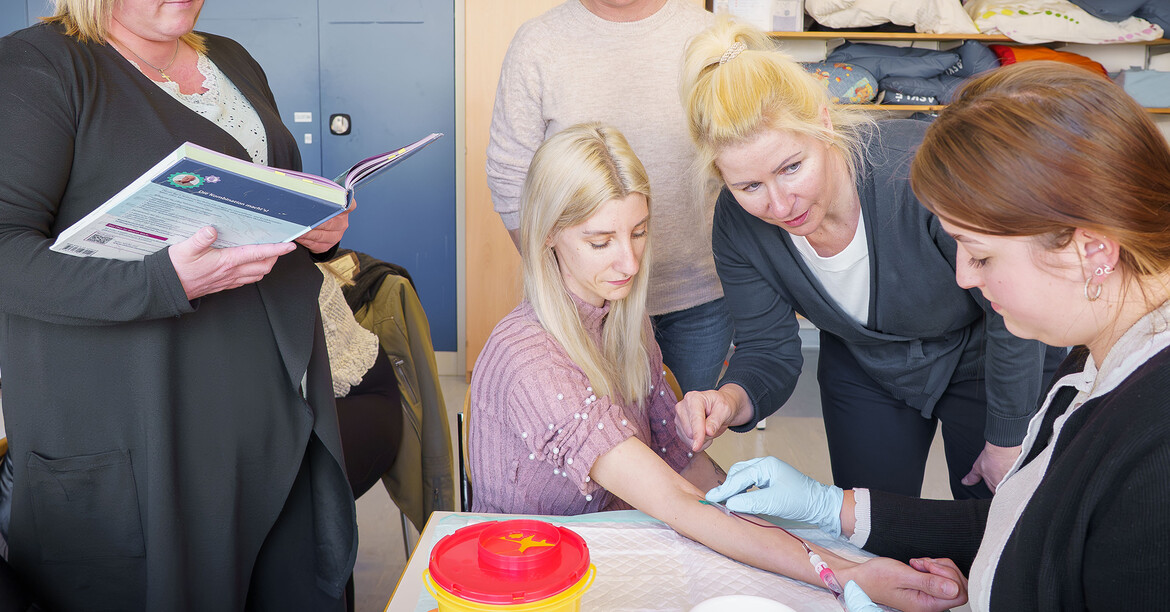 Teilnehmende der SOB-Teilzeitklasse üben unter Anleitung eine Blutabnahme im Schulungsraum.