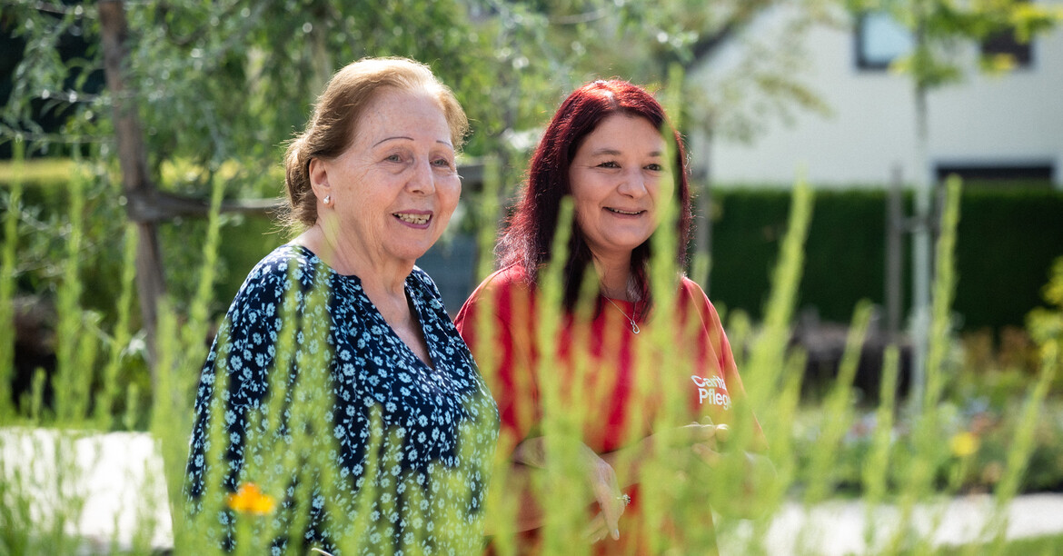 Foto © Karin Wernig, Stadt Villach © Karin Wernig, Stadt Villach Eine ältere Dame und eine Pflegekraft lächeln. Vor ihnen sieht man unscharf grüne Pflanzen wachsen.