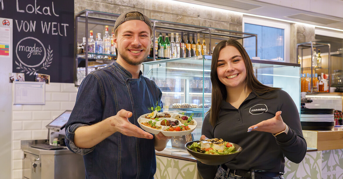 Ein Koch des magdas LOKAL hält einen Mezze-Teller eine Servicekraft einen magdas-Salat, während sie vor der Bar stehen.