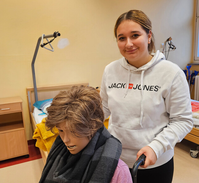 Eine Schülerin lächelt, während sie eine medizinische Übungspuppe im Rollstuhl schiebt.