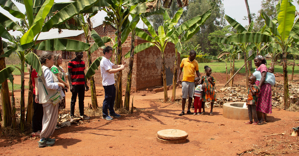 Caritasdirektor Ernst Sandriesser steht mit einer afrikanischen Familie bei den Biogasanlagen. Um sie herum stehen Bananenbäume.