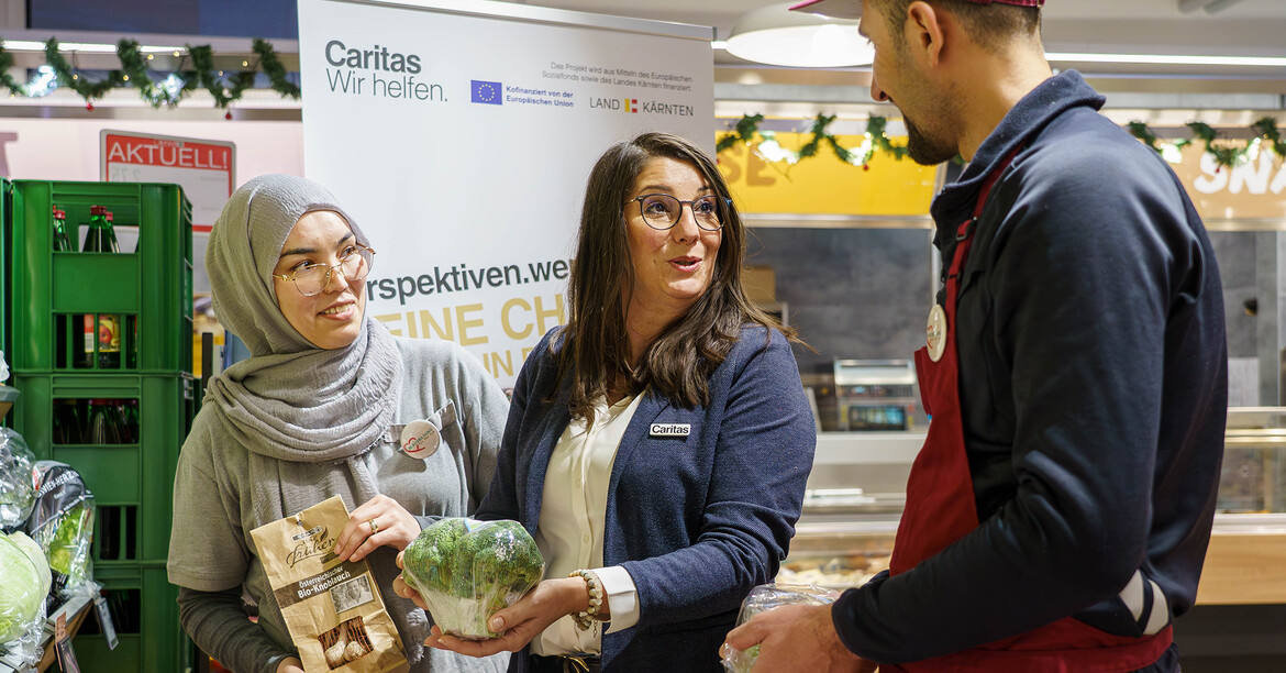 Die Projektleitung des perspektiven.werks steht mit einem Teilnehmer und einer Teilnehmerin bei der Gemüse- und Obstabteilung des SPAR Marktes und sie sprechen miteinander.