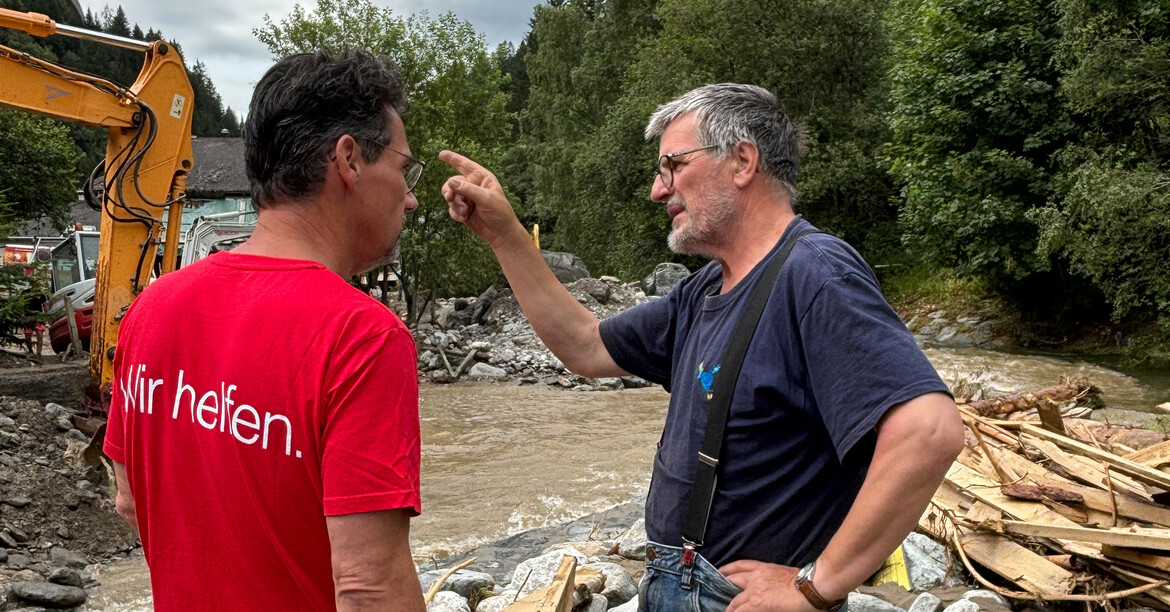 Caritasdirektor Ernst Sandriesser steht vor Schutt der Unwetterkatastrophe und spricht mit einem Betroffenen.