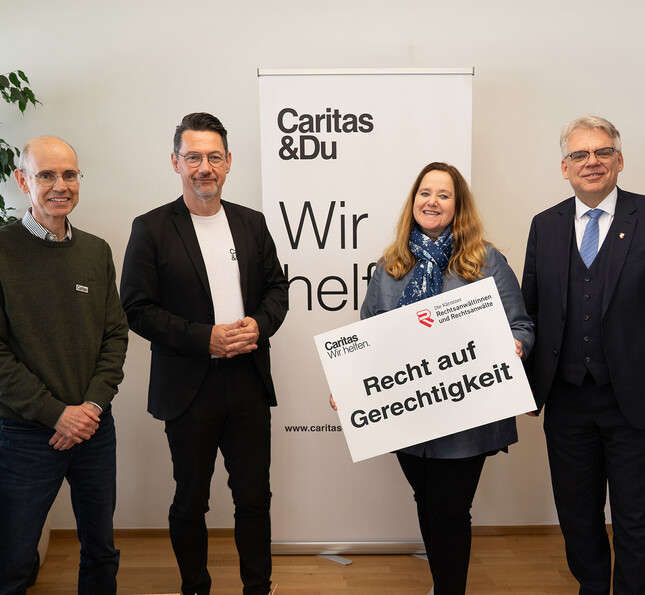 Vier erwachsene Personen stehen in einem Innenraum vor einem Roll-up mit der Aufschrift „Caritas & Du – Wir helfen“. Eine Person hält eine Tafel mit dem Text „Recht auf Gerechtigkeit“.