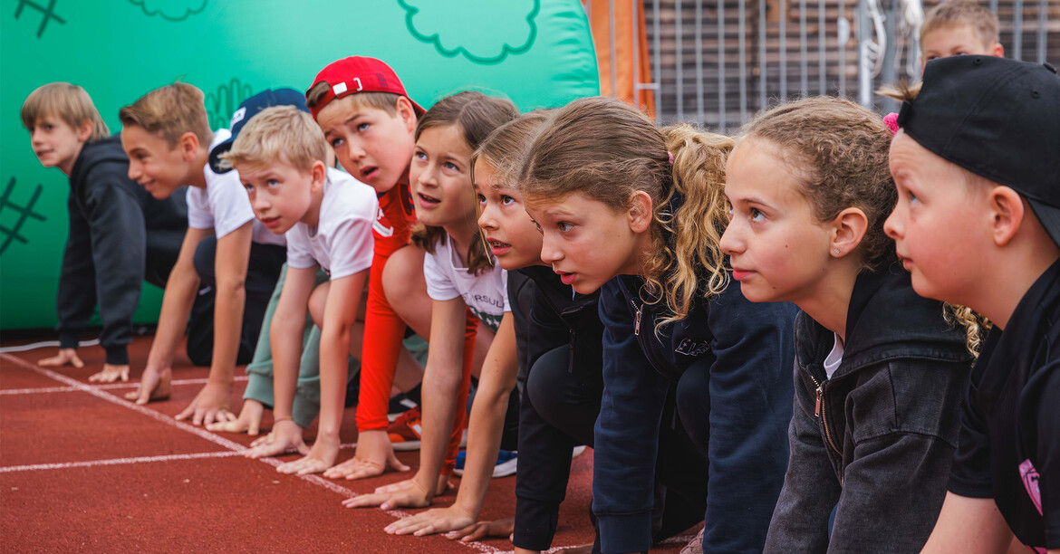 Kinder knien nebeneinander an der Startlinie einer Laufbahn und bereiten sich auf den Lauf vor.