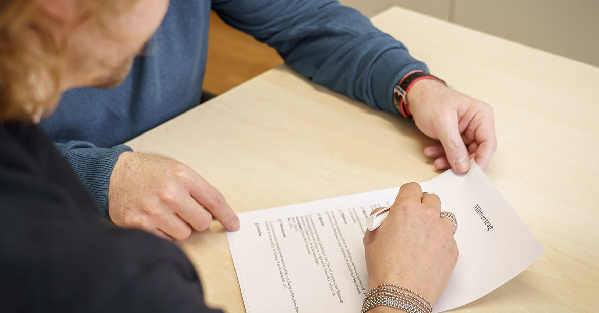 wei Personen sitzen an einem Tisch und betrachten gemeinsam ein Dokument. 