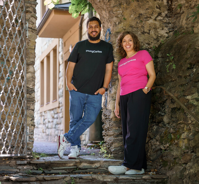 Mario und Theresa, beides youngCaritas-Mitarbeiter*innen, stehen vor einer Steinwand auf einer Stiege und lächeln.