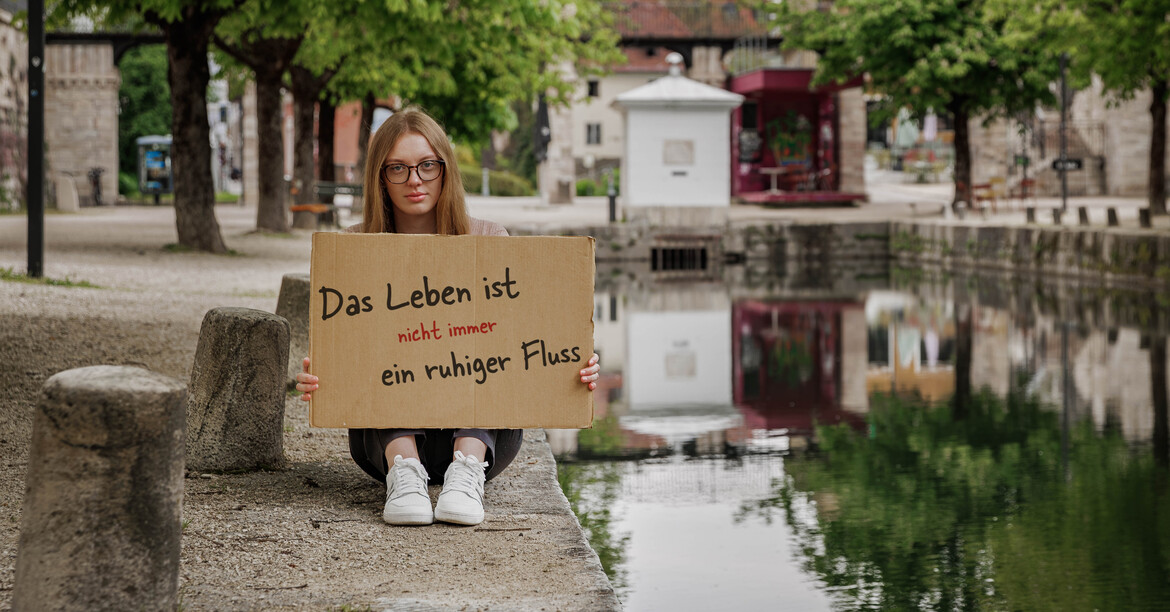 Eine junge Frau sitzt am Rand des Lendkanals in Klagenfurt und hält ein Schild in der Hand "Das Leben ist nicht immer ein ruhiger Fluss".
