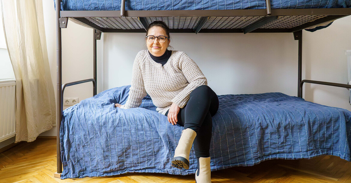 Eine erwachsene Person sitzt auf einem unteren Bett eines Stockbettes in einem einfachen, hellen Zimmer.