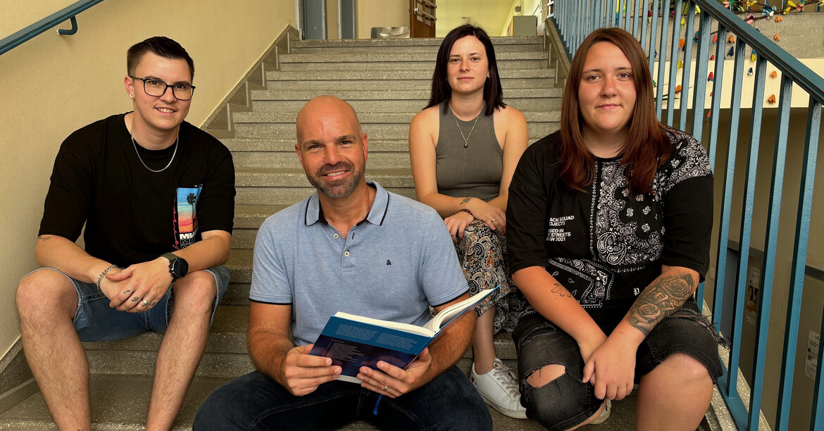 Direktor Wolfgang Kogler sitzt mit drei Schüler*innen auf einer Stiege in der Schule und hält ein offenes Buch in der Hand.