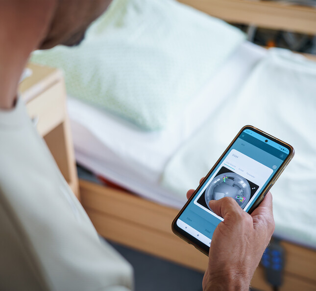 Person bedient eine App zur Raumüberwachung am Smartphone neben einem Pflegebett.