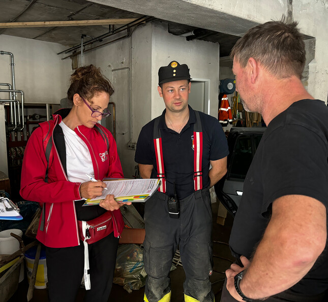 Eine Sozialarbeiterin füllt ein Formular für die Katastrophenhilfe für einen Betroffenen aus. Ein Feuerwehrmann steht neben ihr.
