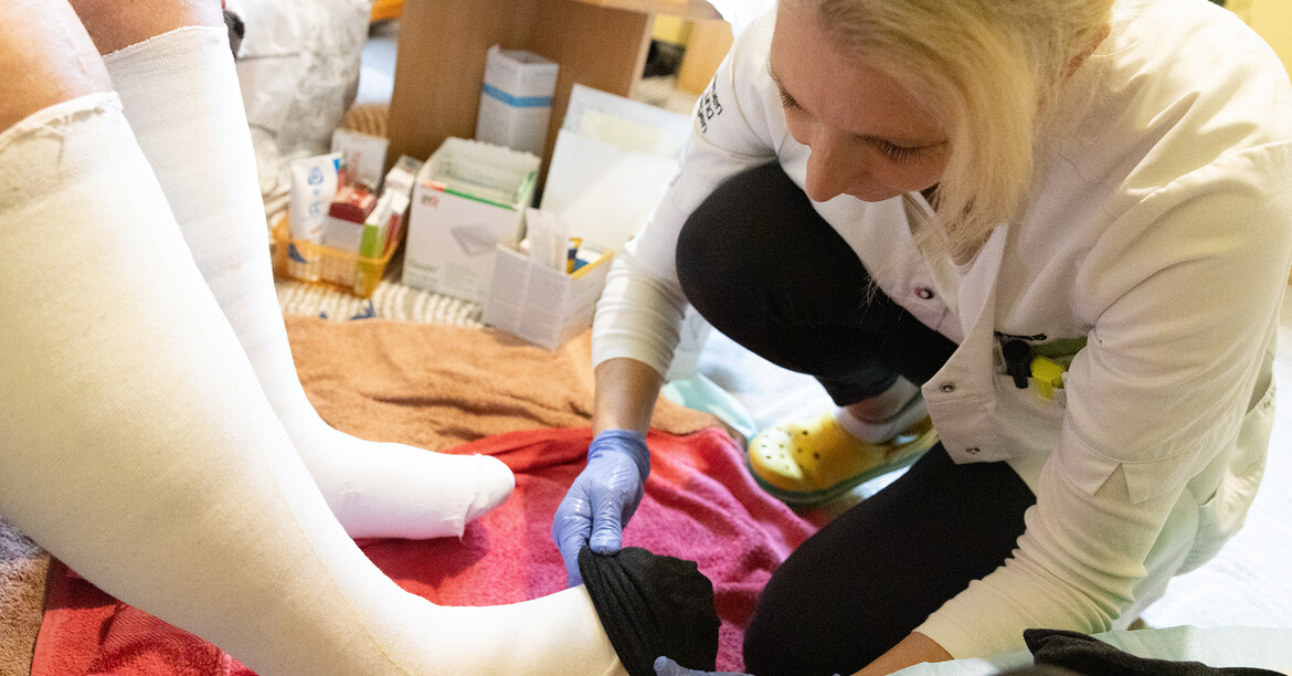 Eine Pflegekraft kniet vor einem Klienten und zieht im Socken über die verbunden Füße an.